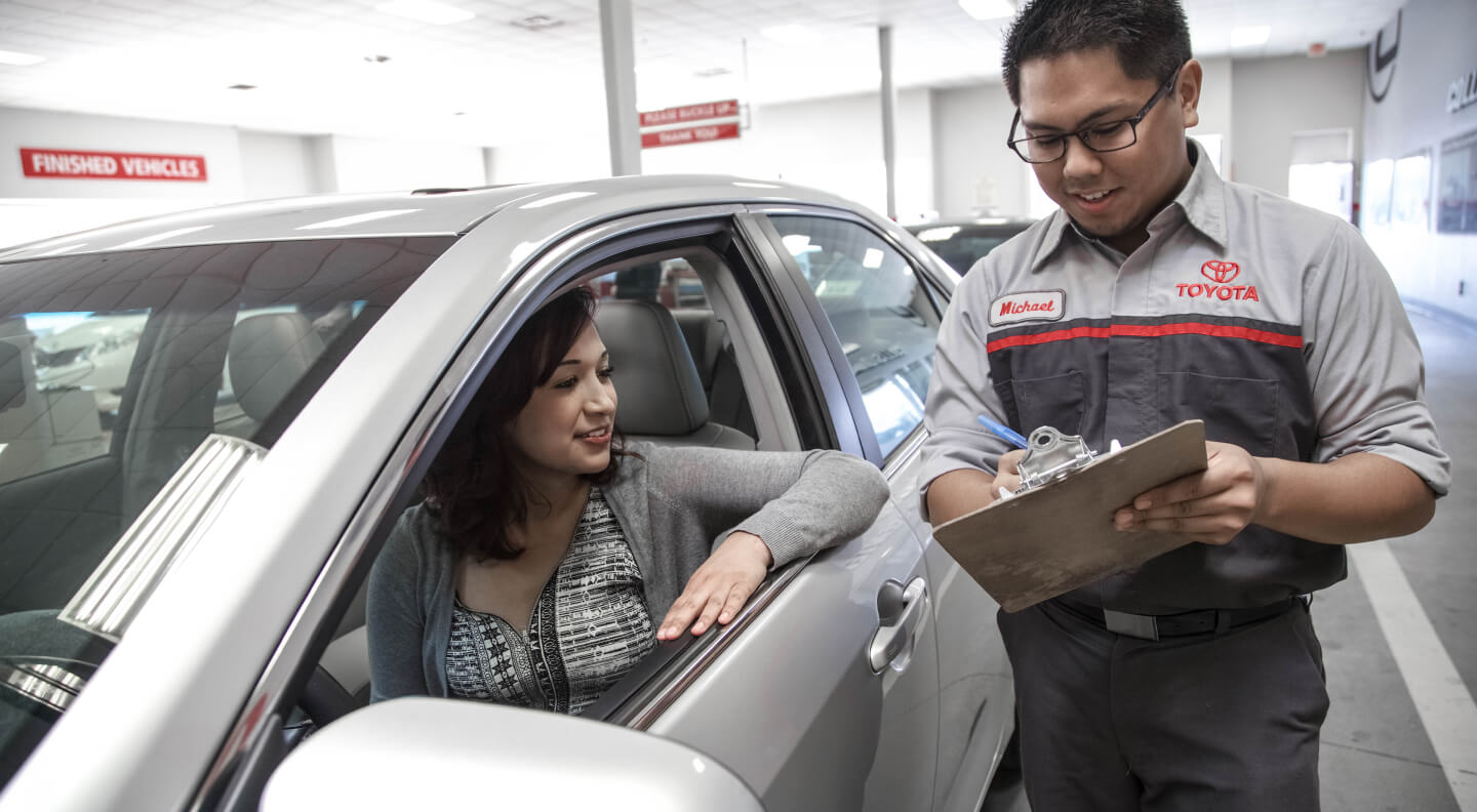 Side view of a Toyota Camry in Silver as a advisor with a clipboard assist a driver.